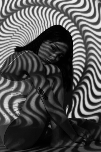 A woman sits near a black and white striped wall, her posture evoking a thinking silhouette—an image that suggests conceptual thought, philosophical reflection, and a sense of inner light
