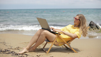 A woman in a yellow shirt sits on a wooden folding chair at the beach, working peacefully by the sea