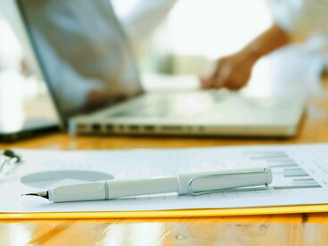 Primo piano di una penna stilografica posata su un documento con grafici, con una persona che lavora al laptop sfocata sullo sfondo