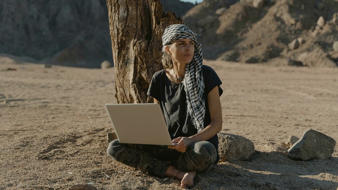 A woman in a black shirt sits on the ground in the desert, working remotely on her laptop
