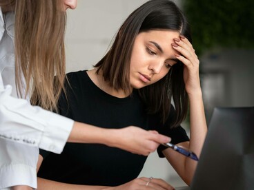 Una giovane donna con un'espressione afflitta guarda il suo laptop, mentre un'altra persona la rimprovera indicando lo schermo con una penna