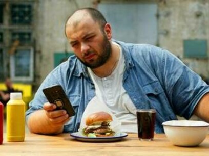 Un uomo siede a un tavolo di legno all'aperto, guardando il suo smartphone. Davanti a lui ci sono un hamburger, un bicchiere di liquido scuro, una ciotola bianca e bottiglie di ketchup e senape: "le combinazioni di carboidrati raffinati e grassi presenti negli alimenti ultra-trasformati producono un “effetto sovra-additivo sui sistemi di ricompensa del cervello”, con tassi di dipendenza ... simili a quelli di sostanze legali come l'alcol o il tabacco"