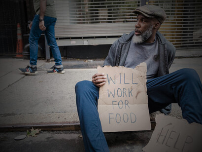 A mature man sits in a doorway holding a cardboard sign, highlighting homelessness and poverty in the US