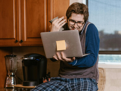 At the kitchen counter, a man talks on the phone and scrolls through news on his computer, feeling excited but not realizing the information is false