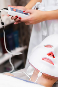 A client relaxes on a cosmetology table in a salon, wearing a white facial mask while enjoying the rejuvenating effects of red light therapy