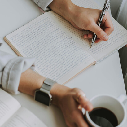 An author writes stories by hand in a notebook, with an open book and pen beside them