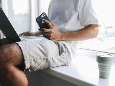 A Gen Z man sits at his desk, scrolling on his phone while working on his laptop — a scene of everyday digital multitaskin