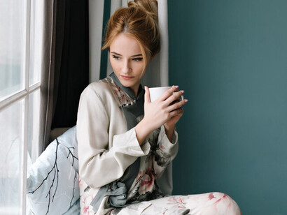 A young blonde woman sipping tea while gazing out of a large window, enjoying the peaceful atmosphere of her home