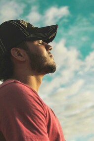 A man in a black cap stands with his eyes closed beneath a cloudy sky, lost in thought