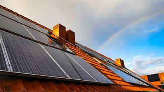 A solar panel mounted on a roof, with a vibrant rainbow arching across the sky in the background