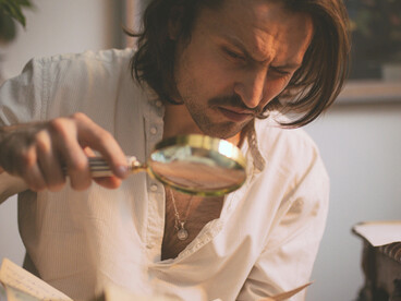 A solitary writer sits at a desk, smoking a cigarette and holding a sheet of paper in a dimly lit room