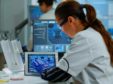 Una scienziata in camice bianco e occhiali protettivi lavora in un laboratorio moderno, utilizzando un tablet per analizzare dati o immagini di strutture biologiche