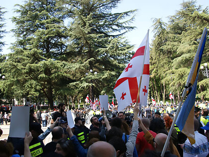 A large crowd of protesters marches through city streets in Georgia, holding national flags while organisers speak through a megaphone