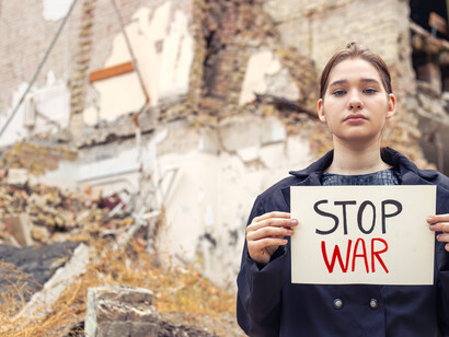A girl standing in the rubble with “stop war,” reflecting the reality behind discussions of expansion, deterrence, and security