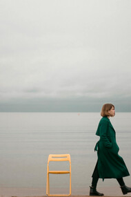 A woman in a green coat walks away from a chair by the ocean