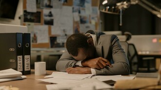 A man in a gray long-sleeved shirt rests his head on the table, crying at work