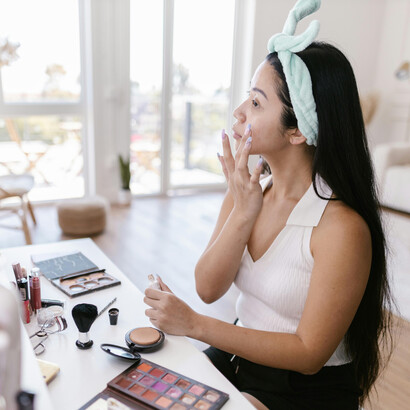 A long-haired brunette applies makeup in a brightly lit room