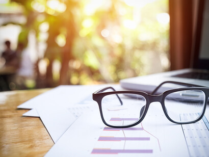 Occhiali da vista appoggiati su documenti e grafici su una scrivania di legno, con un laptop sullo sfondo