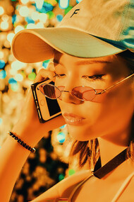 A girl wearing sunglasses and a cap, talking on the phone in front of neon lights