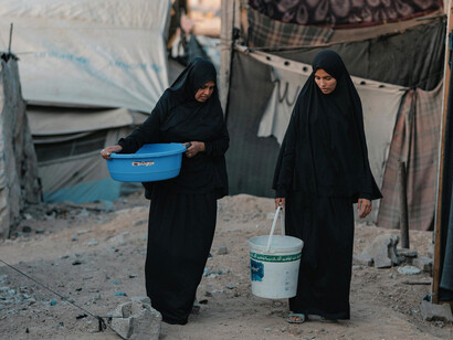 Women carrying water in a camp, representing the human cost of systemic underdevelopment