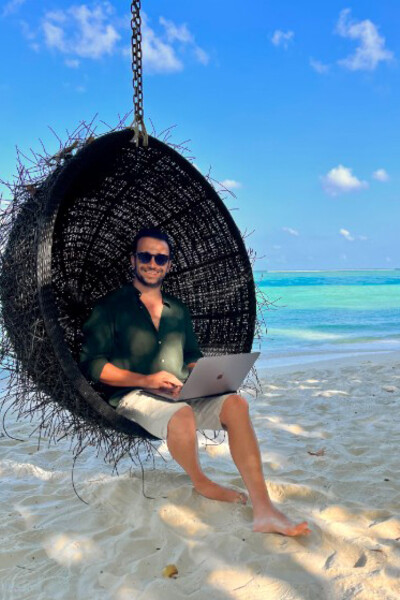 A freelancer working on his laptop while sitting in a hanging chair on a beach in the Maldives