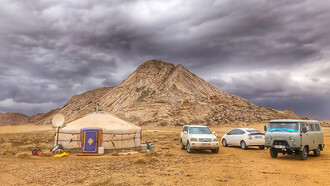 Three vehicles parked beside a yurt in Bayan-Unjuul, Töv, Mongolia, with mountains looming under a cloudy sky