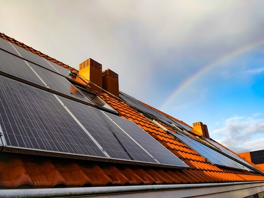 A solar panel mounted on a roof, with a vibrant rainbow arching across the sky in the background
