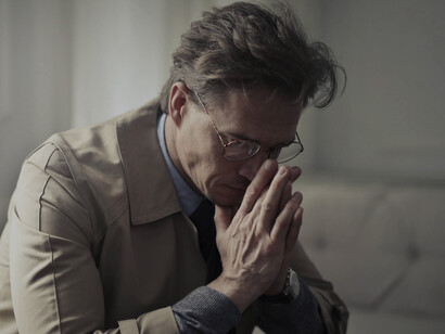 A mature man sits on a couch in a dimly lit living room, thinking alone