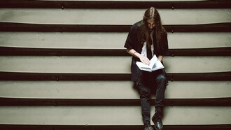 A person sitting on a set of stairs, holding a book, lost in thought or overwhelmed by the weight of their studies
