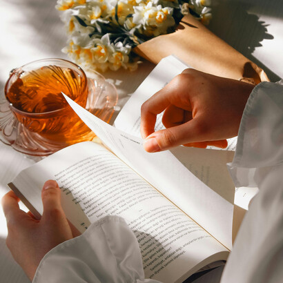 A woman rests with a book in her hands, a cup of tea and a bouquet of daffodils softly waiting by her side