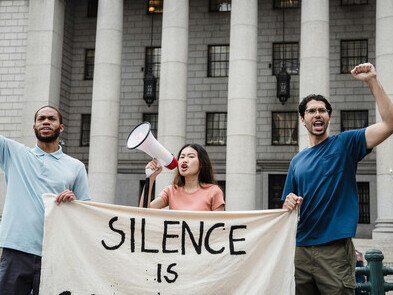 A group of people are protesting on the street, holding a sign that reads “Silence is compliance”