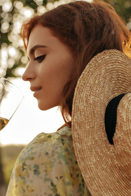 Stylish young girl with a foxy hairstyle donning a modern straw hat and trendy summer outfit poses elegantly with a glass of white wine amidst the picturesque vineyards of Hungary