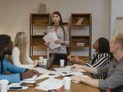 A professional woman in a gray blazer shares her report at a board meeting, engaging in strategic brainstorming with colleagues
