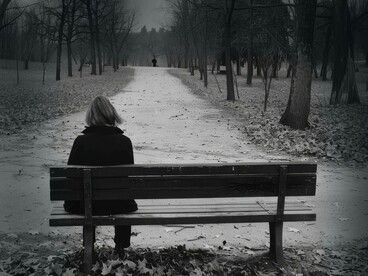 Persona seduta da sola su una panchina in un parco, con l'ambiente circostante spoglio e un'atmosfera generale cupa