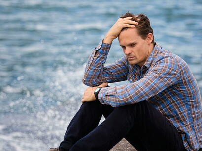 Wearing a blue and brown plaid shirt, the man touches his hair while appearing depressed and alone