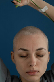 A shaved young woman with eyes closed and arms reaching up