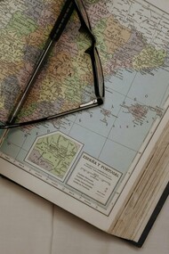 A pair of glasses rests on an open map, alongside a passport from the Bolivarian Republic of Venezuela, visually capturing the forced journeys and global scattering of Venezuelan exiles as they navigate new lives far from their homeland