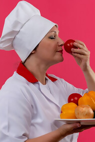 A middle-aged female chef in a chef's uniform, holding a plate of vegetables, sniffing a tomato in her hand