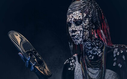 A person from an indigenous African tribe wearing traditional makeup and costume while staring at a mask in their hand, symbolizing the struggle of the modern African youth caught between tradition and modernity
