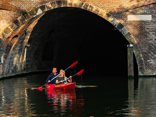 A man and woman paddle a red kayak along a river in Utrecht, the Netherlands, under the bright daytime sky