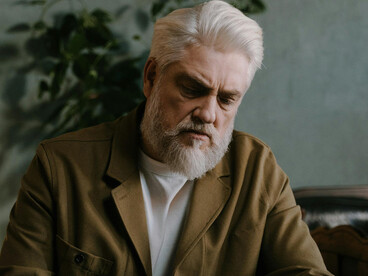 An elderly man sits thoughtfully at a table, holding a pen as he reflects and writes