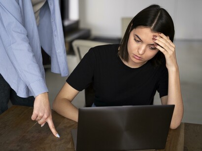 Una ragazza è seduta al suo laptop con la testa tra le mani, mentre un'altra persona si china su di lei e le indica qualcosa sullo schermo, creando un'atmosfera di tensione 