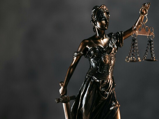 A sculpted defender of law with suspended scales, representing the fragile balance of democratic institutions
