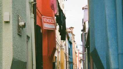 Un vicolo stretto a Burano, Italia, fiancheggiato da edifici dai colori vivaci, con un cartello rosso con la scritta "Vendesi" su uno degli edifici