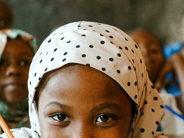 An African schoolgirl smiles while writing with a pencil in class, as fellow students study in the background, symbolizing academic pursuit and childhood education