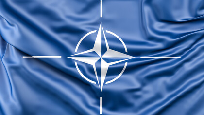 The NATO symbol resting on blue satin, conveying how global order is often staged as harmony while driven by strategic profit