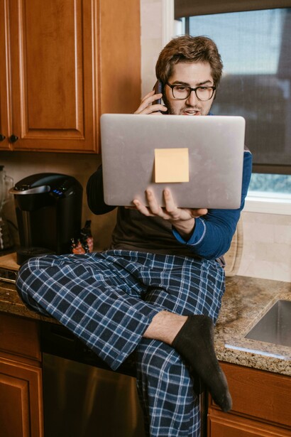 A man stands at his kitchen counter, talking on the phone and checking the news on his computer, excited by what he sees, unaware that it is fake news