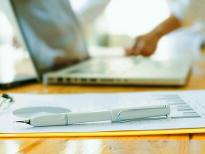 Primo piano di una penna stilografica posata su un documento con grafici, con una persona che lavora al laptop sfocata sullo sfondo