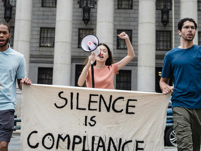 A crowd gathers in protest, holding up a sign with the message “Silence is compliance”