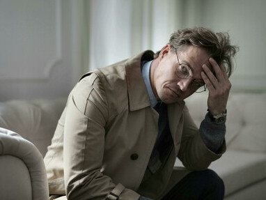 A man in a brown jacket rests on a couch, his expression filled with melancholy